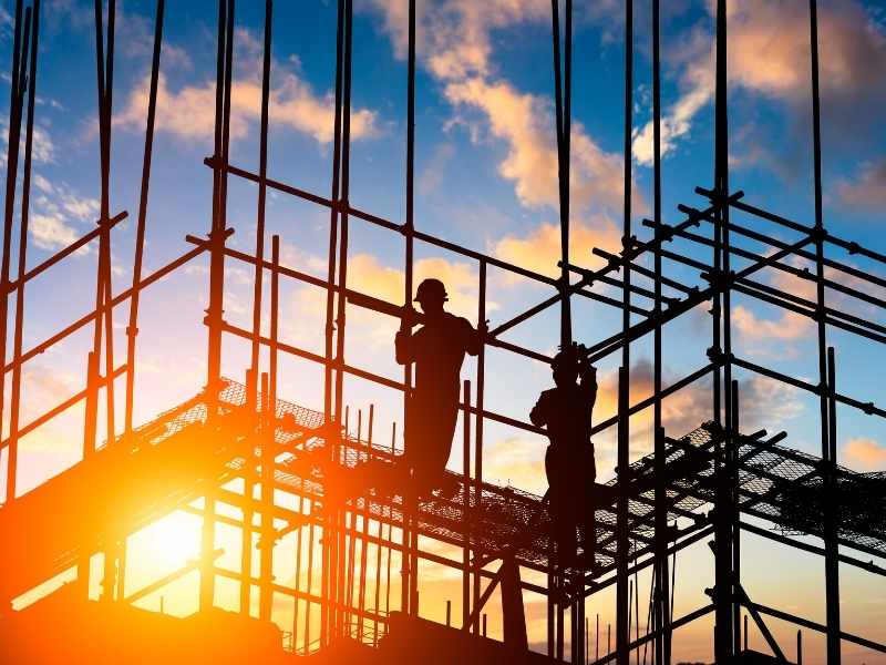 Primer plano de un plano arquitectónico sobre una mesa de obra, con trabajadores al fondo en una estructura de acero. Estética industrial y enfocada.