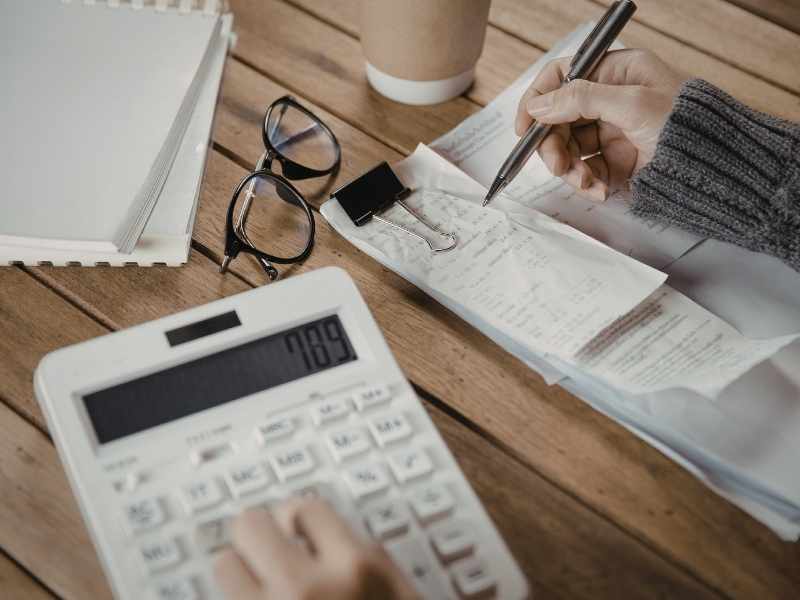 Primer plano de una persona utilizando una calculadora blanca y tomando notas sobre recibos y facturas, representando el control de costos en las finanzas corporativas.